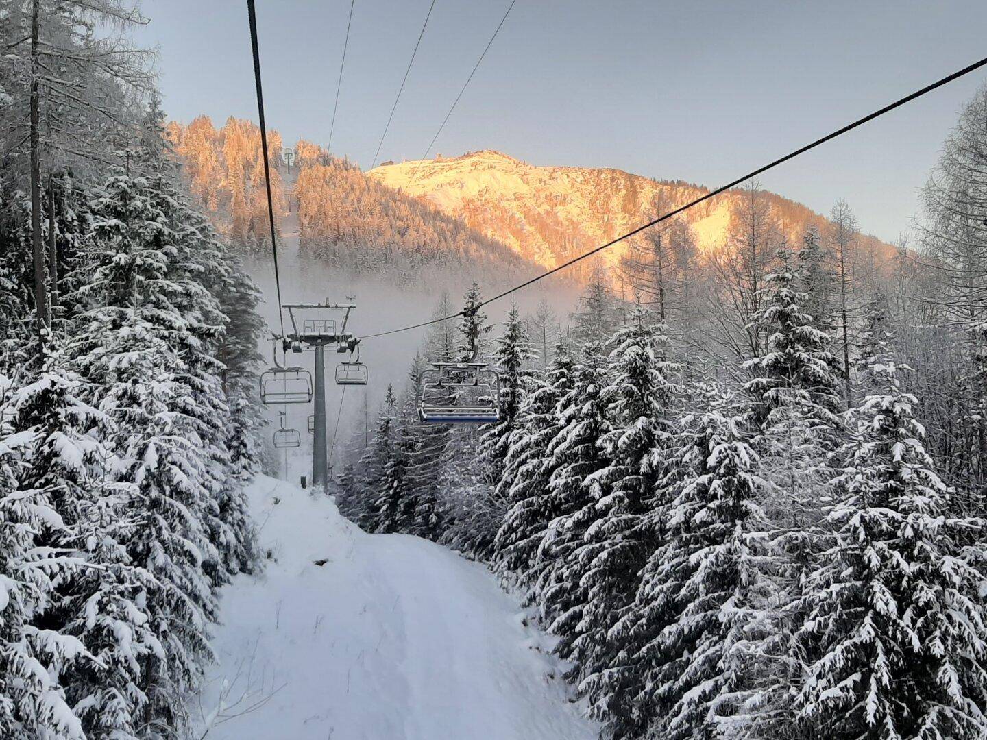 Skilift über einer verschneiten Waldlandschaft mit Bergen im Hintergrund bei Sonnenaufgang.