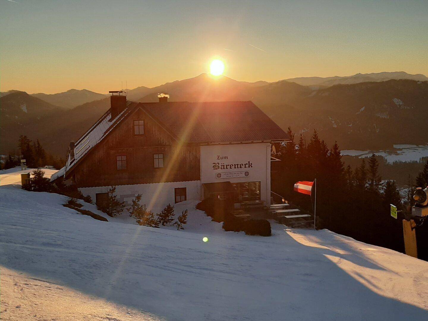 Ein traditionelles Gebäude mit Holzfassade in einer verschneiten Berglandschaft bei Sonnenuntergang, mit österreichischer Flagge.
