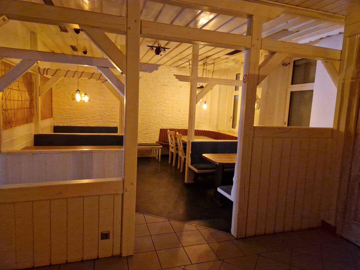 Innenraum mit rustikaler Holzbalkenstruktur und Sitzbänken, beleuchtet von warmen Hängelampen.