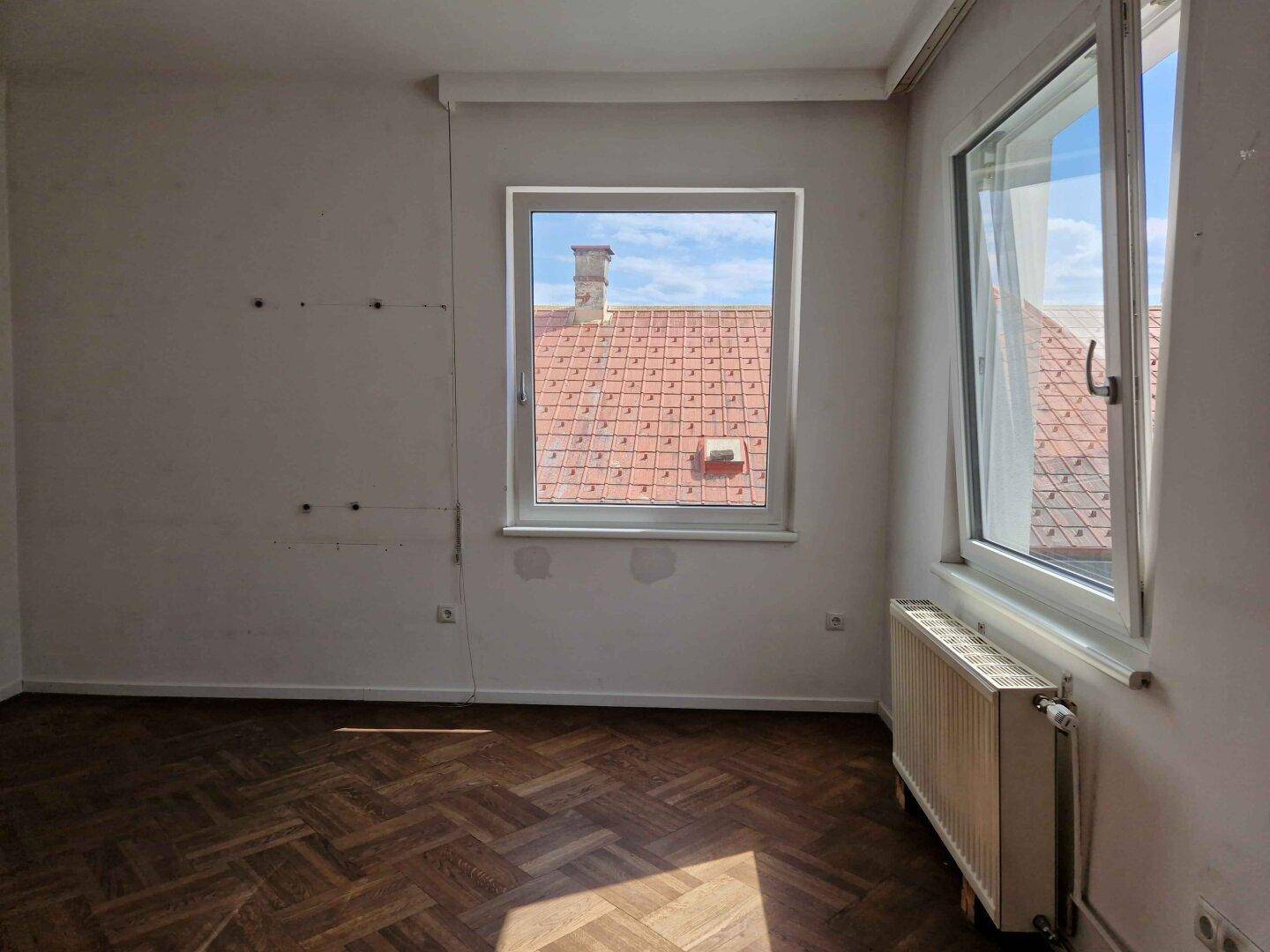 Geräumiges Zimmer mit Parkettboden und Blick auf die Dächer und die grüne Umgebung.