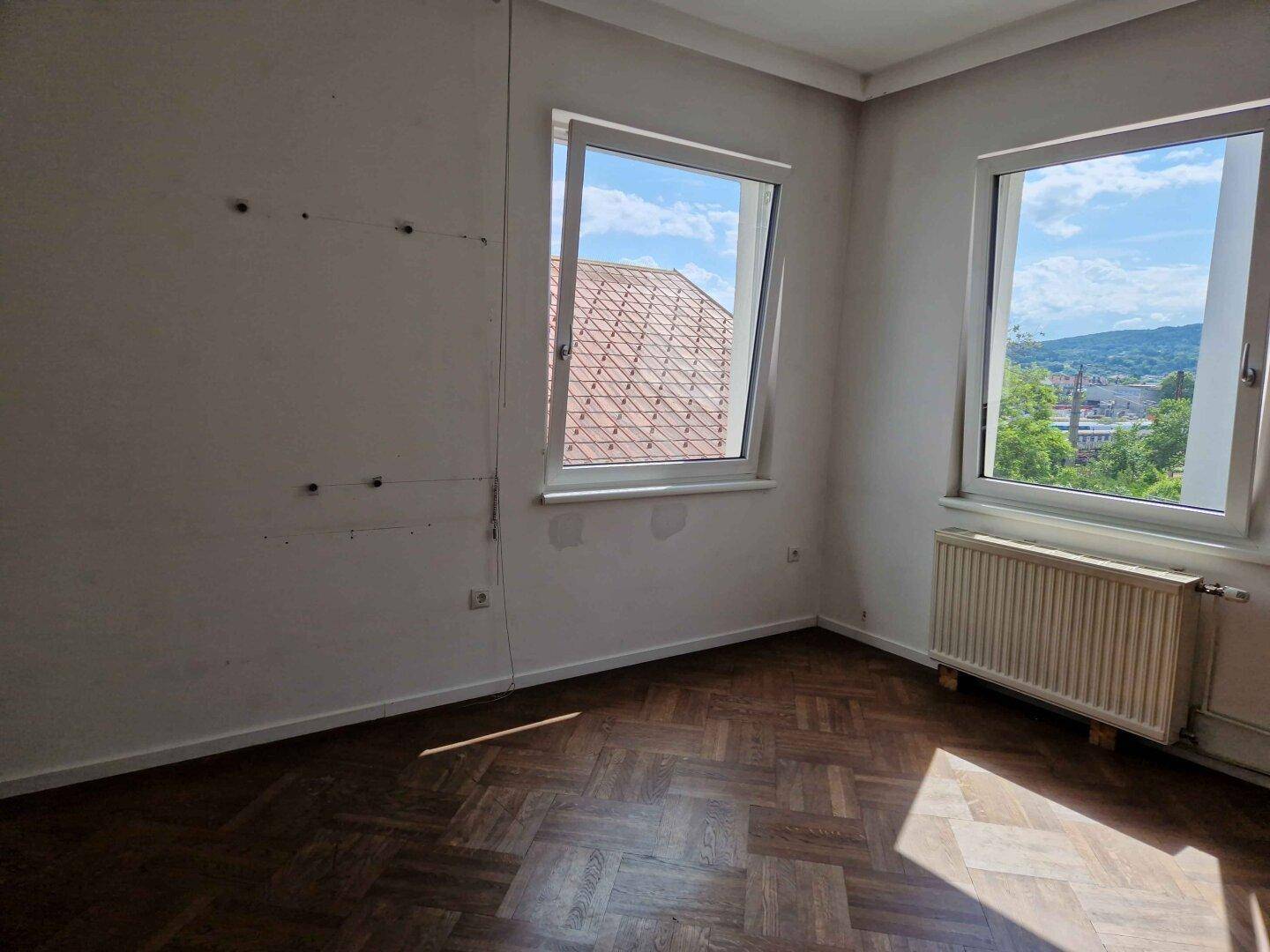 Helles Zimmer mit Fischgrätparkett und zwei Fenstern, die einen weiten Ausblick bieten.