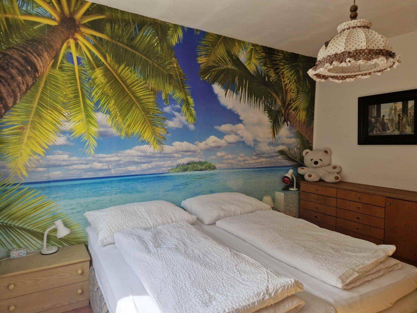 Schlafzimmer mit Doppelbett, Nachttischen und einer lebhaften Wandtapete mit Palmenmotiv.