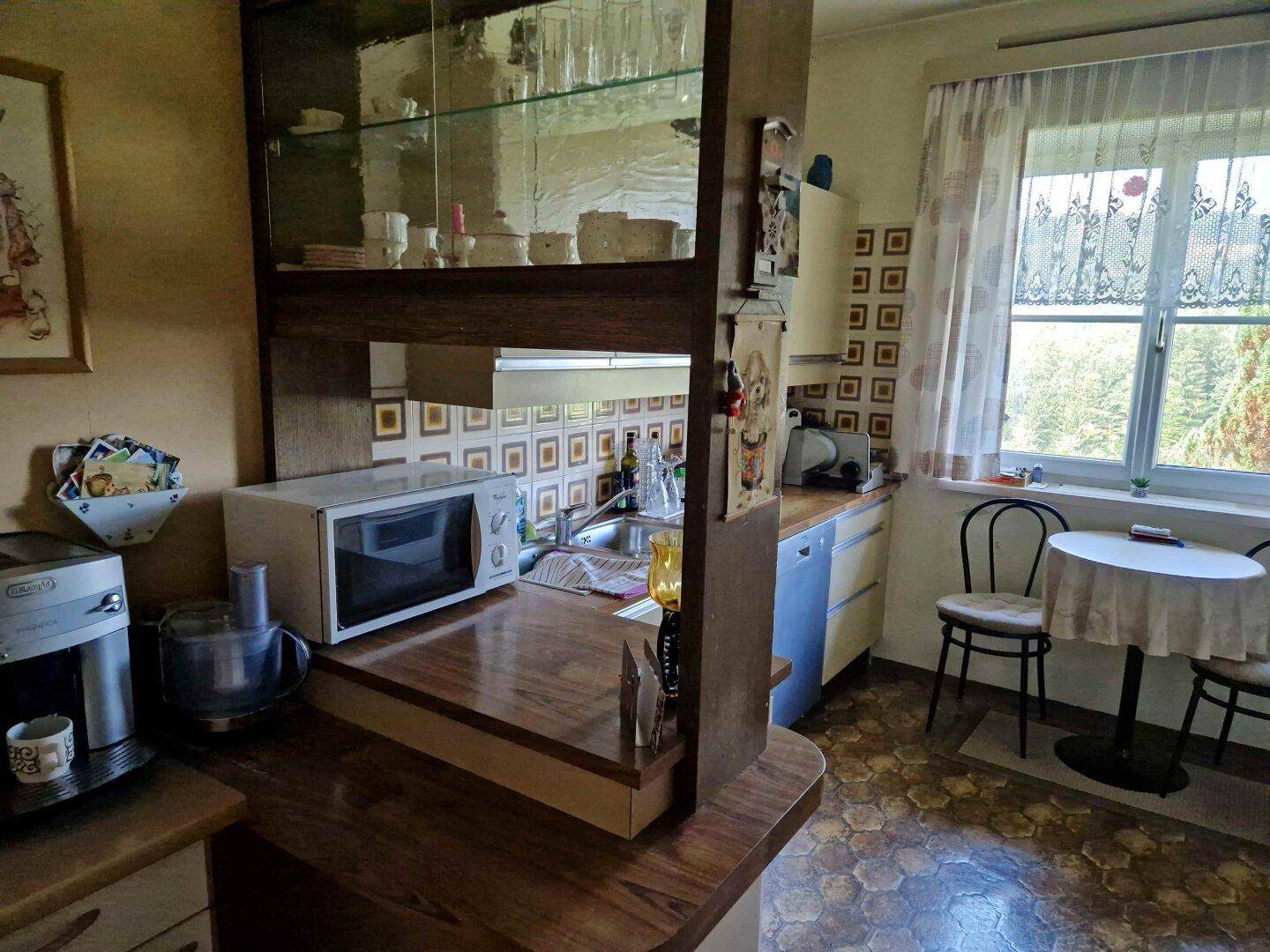 Funktionale Küche mit Frühstücksbar, älteren Geräten und einem kleinen Essbereich am Fenster mit Blick ins Grüne.