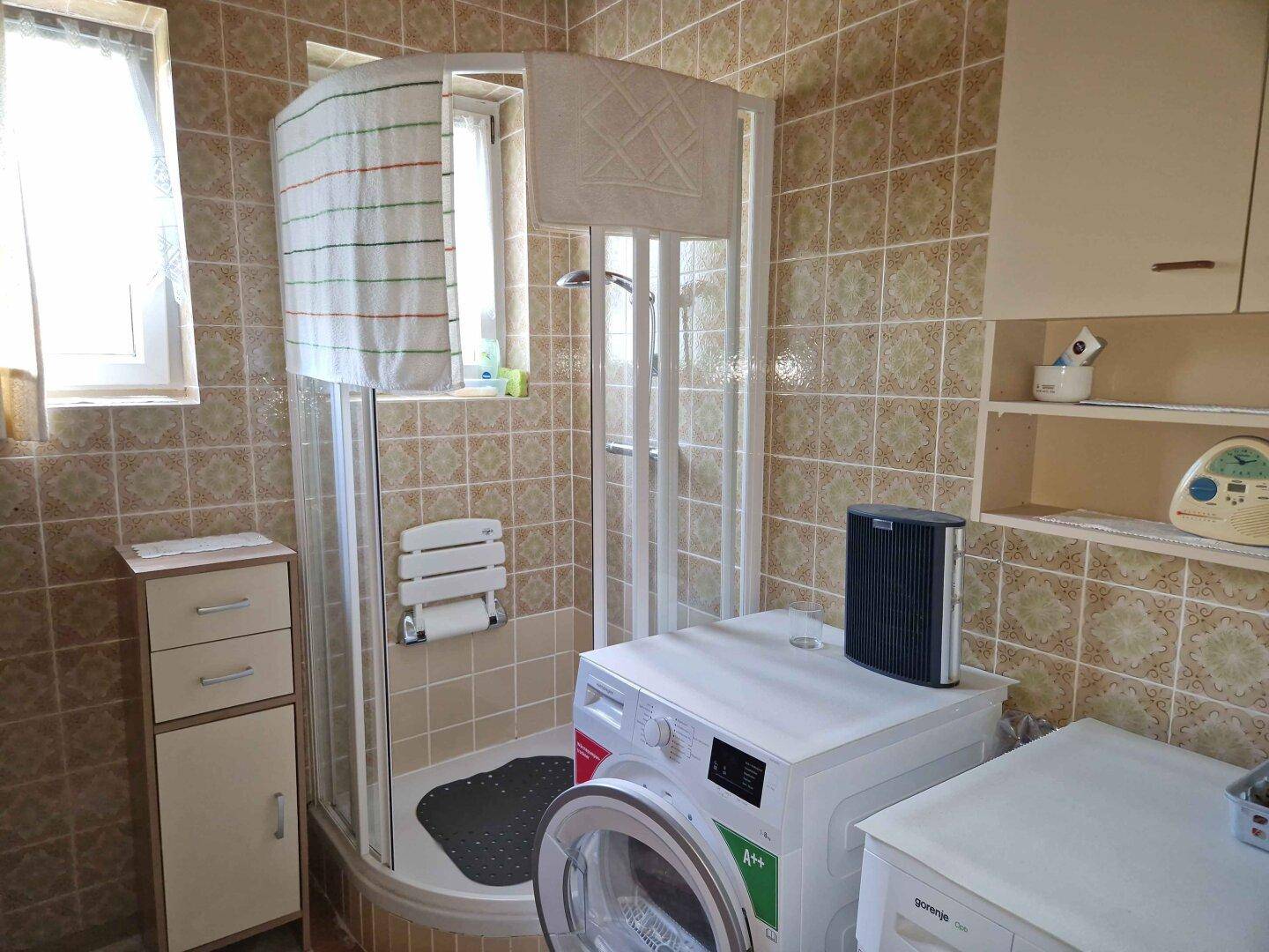 Gefliestes Badezimmer mit Duschkabine, Waschmaschine und Fenster, funktional und hell gestaltet.