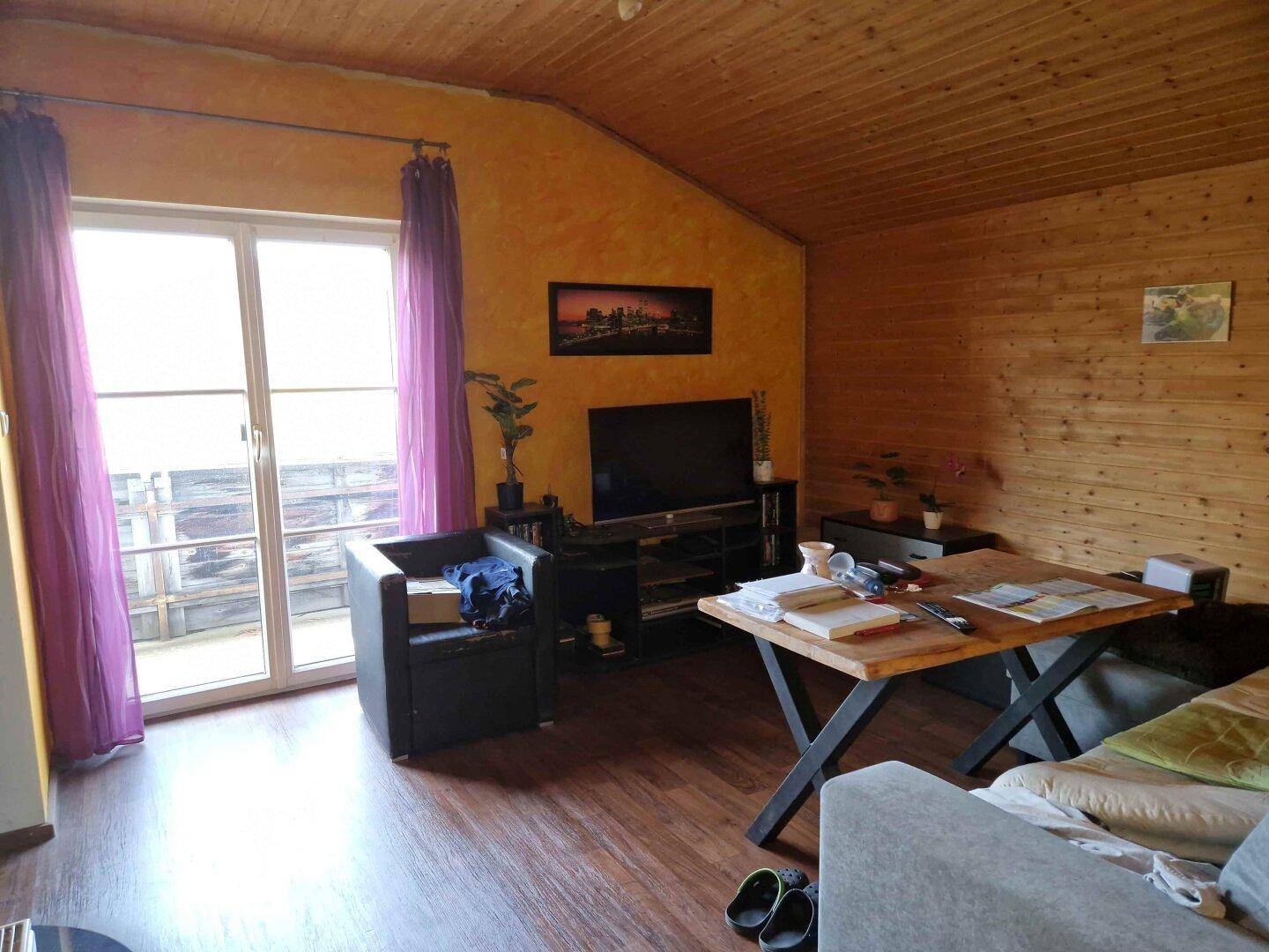Rustikales Wohnzimmer mit Holzverkleidung an Wänden und Decke, Zugang zu einem Balkon und viel Tageslicht.