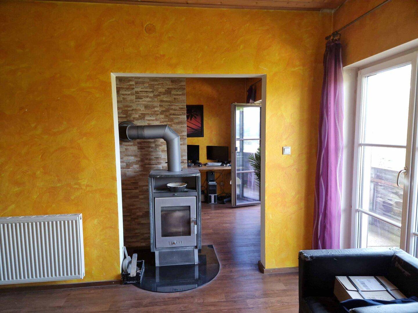 Wohnzimmer mit gemütlichem Holzofen und offenem Durchgang zu einem weiteren Raum, warme Atmosphäre.