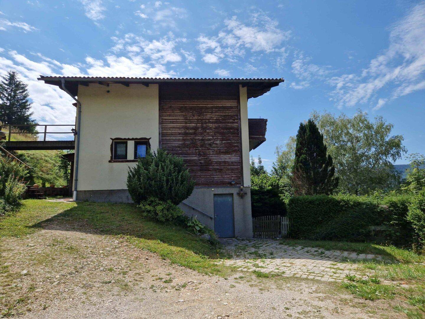 Seitenansicht des Hauses mit Zugang zum Keller und einem Schotterweg, umgeben von Grünflächen.