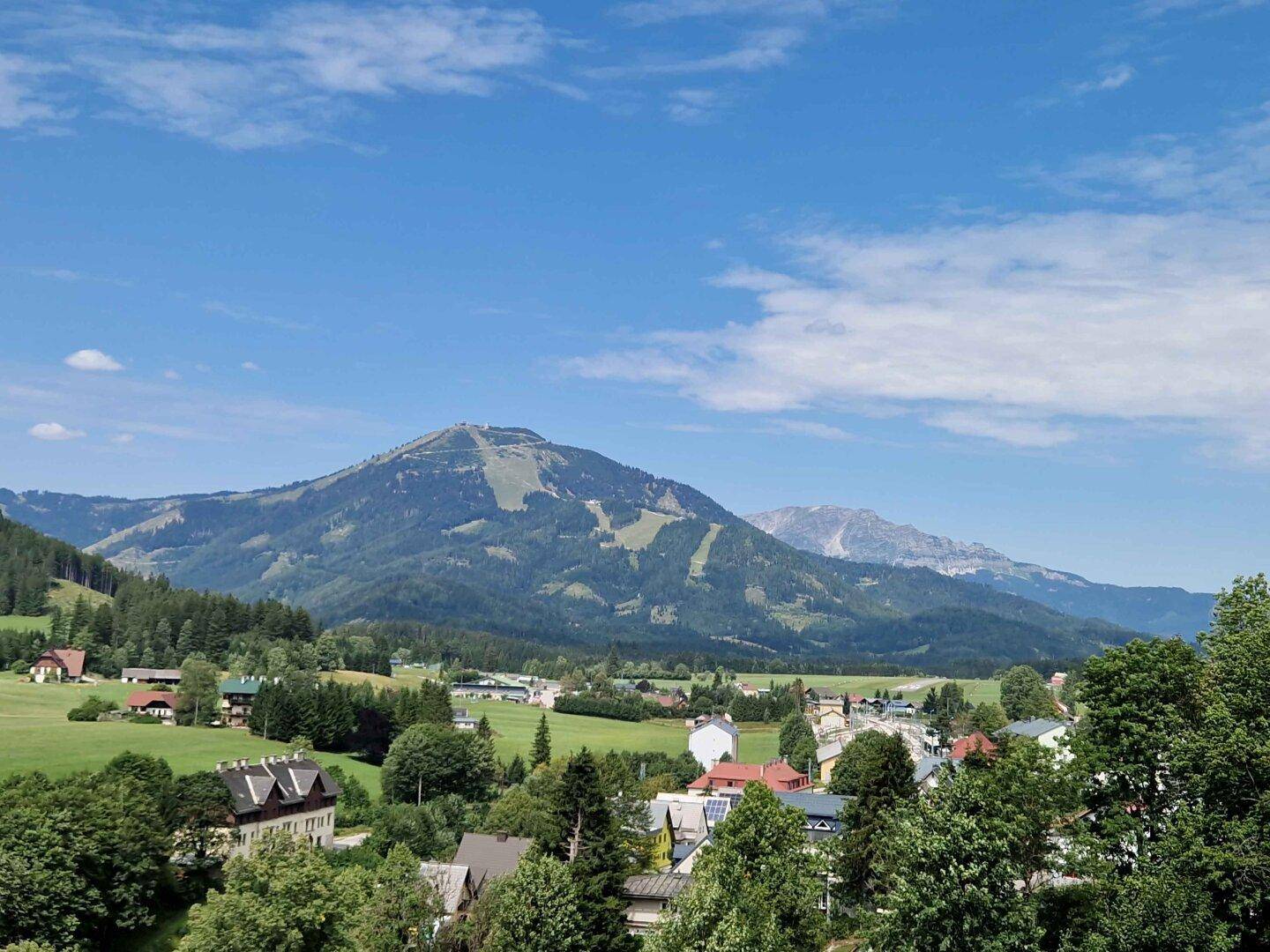 Panoramablick auf ein idyllisches Dorf im Tal, umgeben von beeindruckenden Bergen und grünen Hügeln.