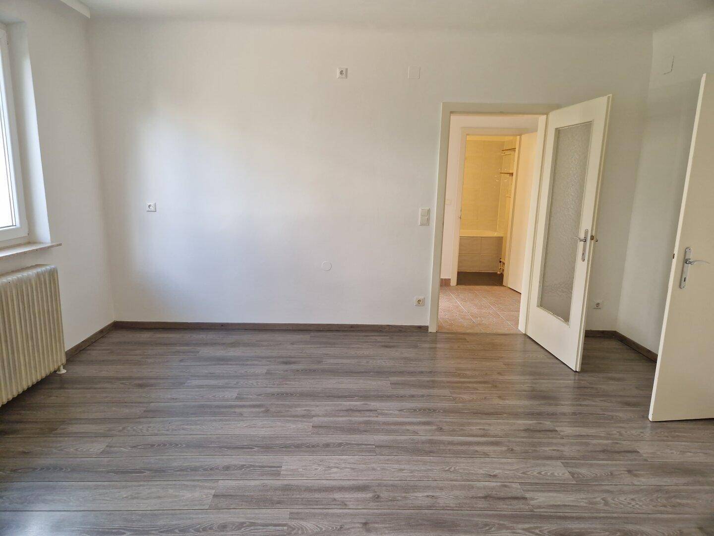 Geräumiger, leerer Raum mit Laminatboden und einer Tür, die zu einem weiteren Zimmer führt.