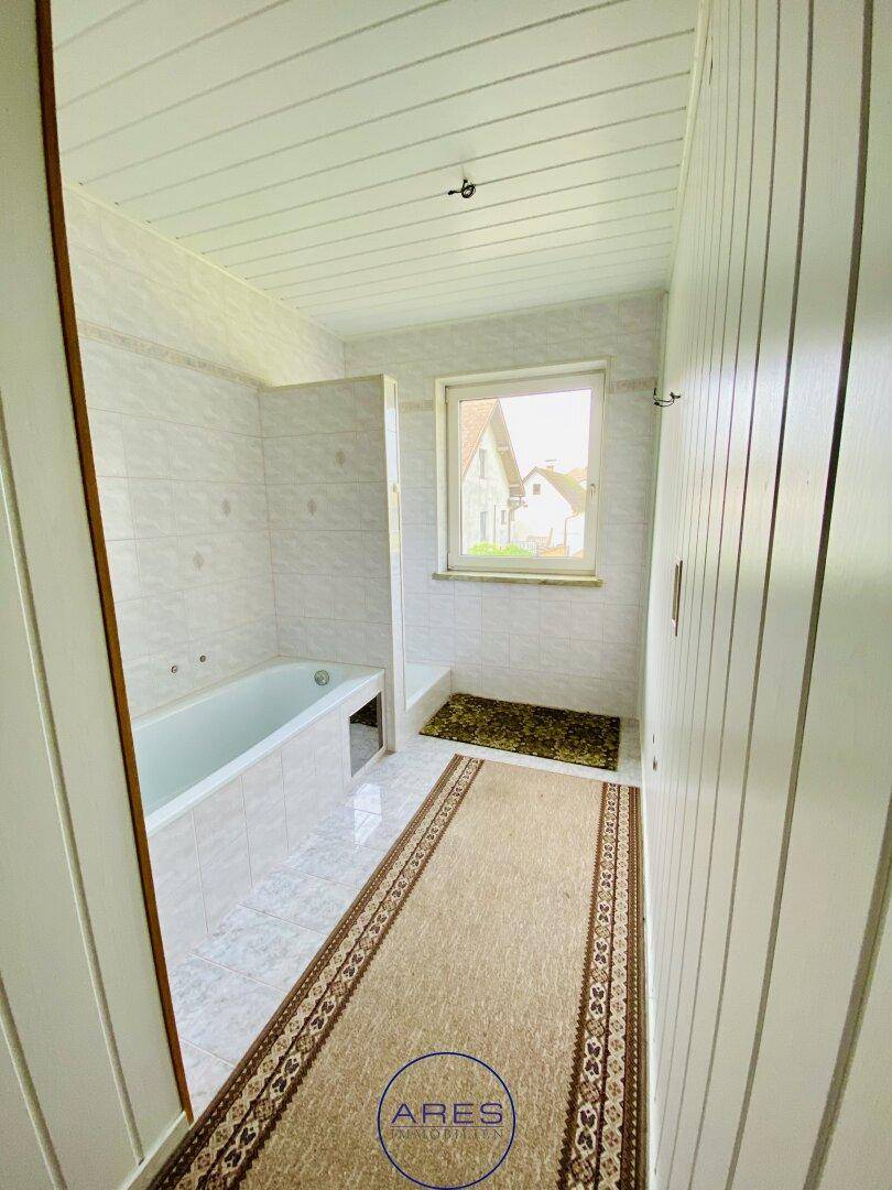 Badezimmer mit Badewanne, hellen Fliesen, Fenster und einem Teppich auf dem Boden.