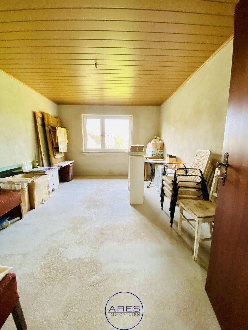 Leerer Raum mit hellen Wänden, Holzdeckenverkleidung und einem Fenster, einige Stühle gestapelt.