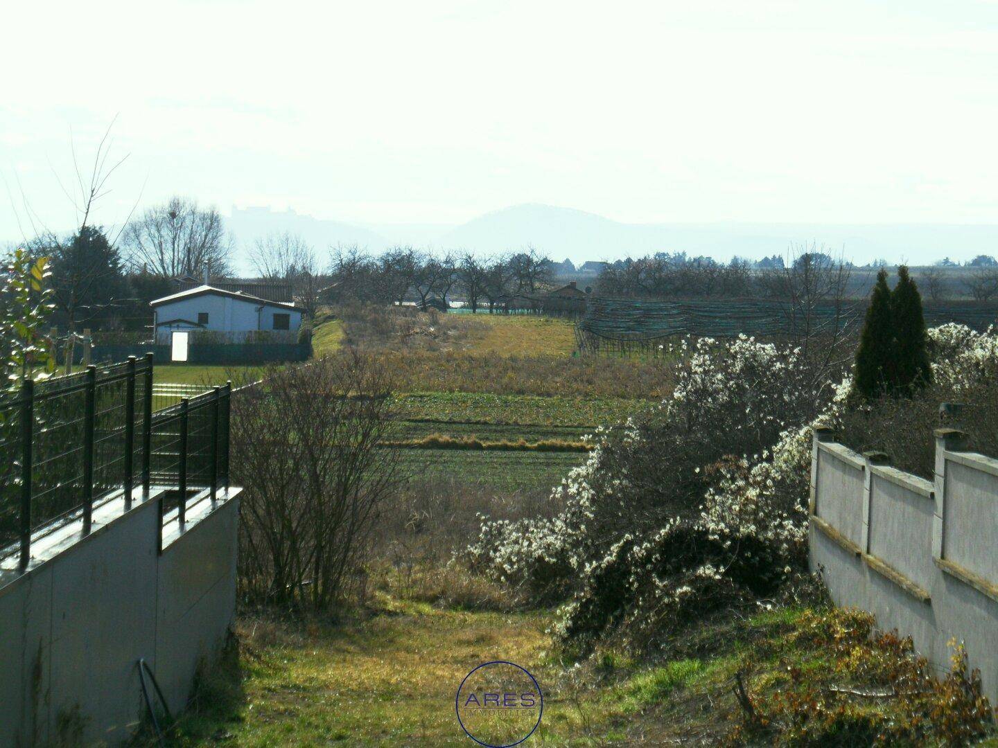 Blick von erhöhter Position über Felder und entfernte Häuser, umrahmt von einer modernen Struktur und Mauer.