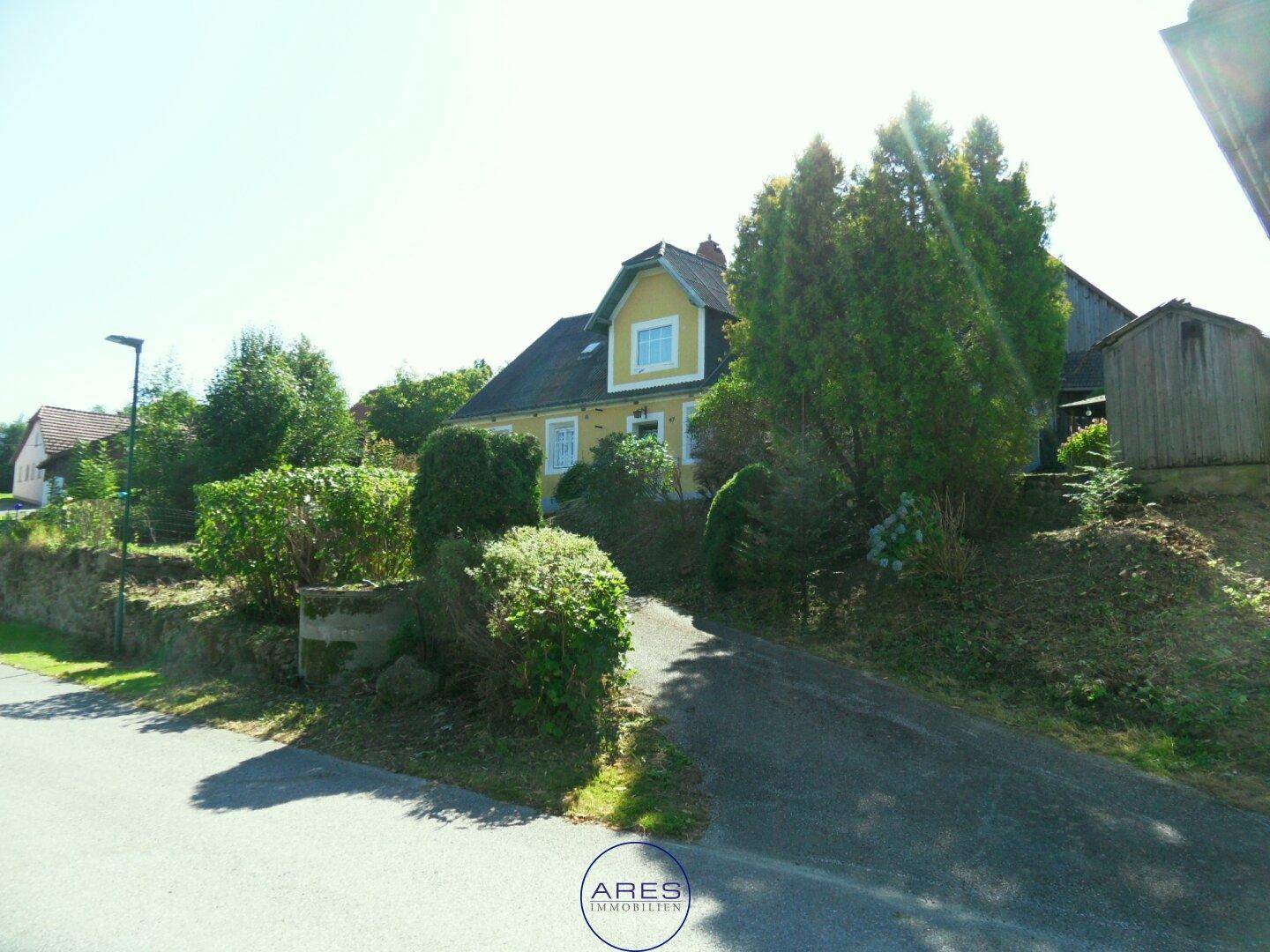 Gelbes Haus mit Zufahrt und üppiger Bepflanzung in ländlicher Umgebung an einem sonnigen Tag.