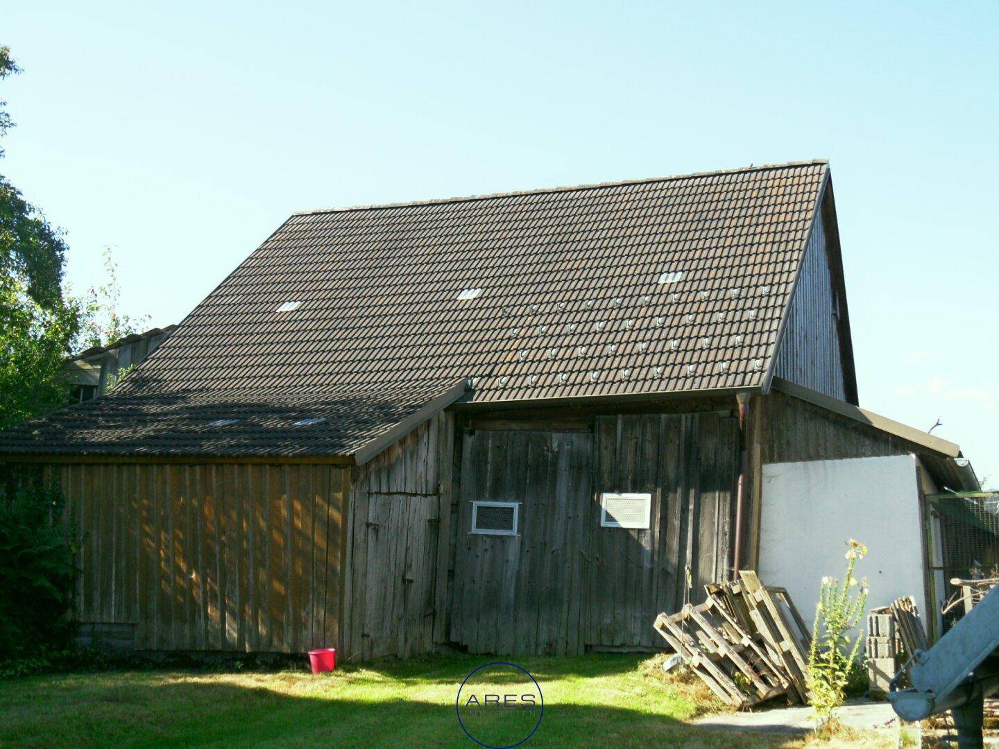 Rustikales Holzgebäude mit Ziegeldach und gestapelten Paletten auf einer grünen Wiese.