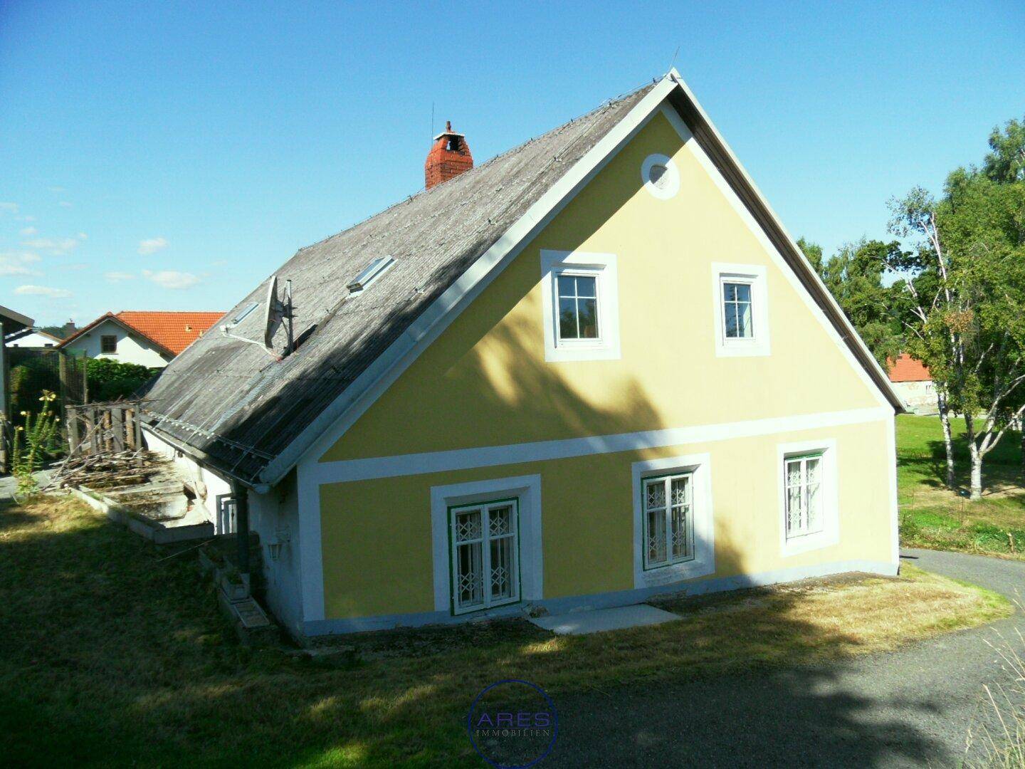 Rückansicht des gelben Hauses mit schrägem Dach und Fenstern, umgeben von grüner Natur.