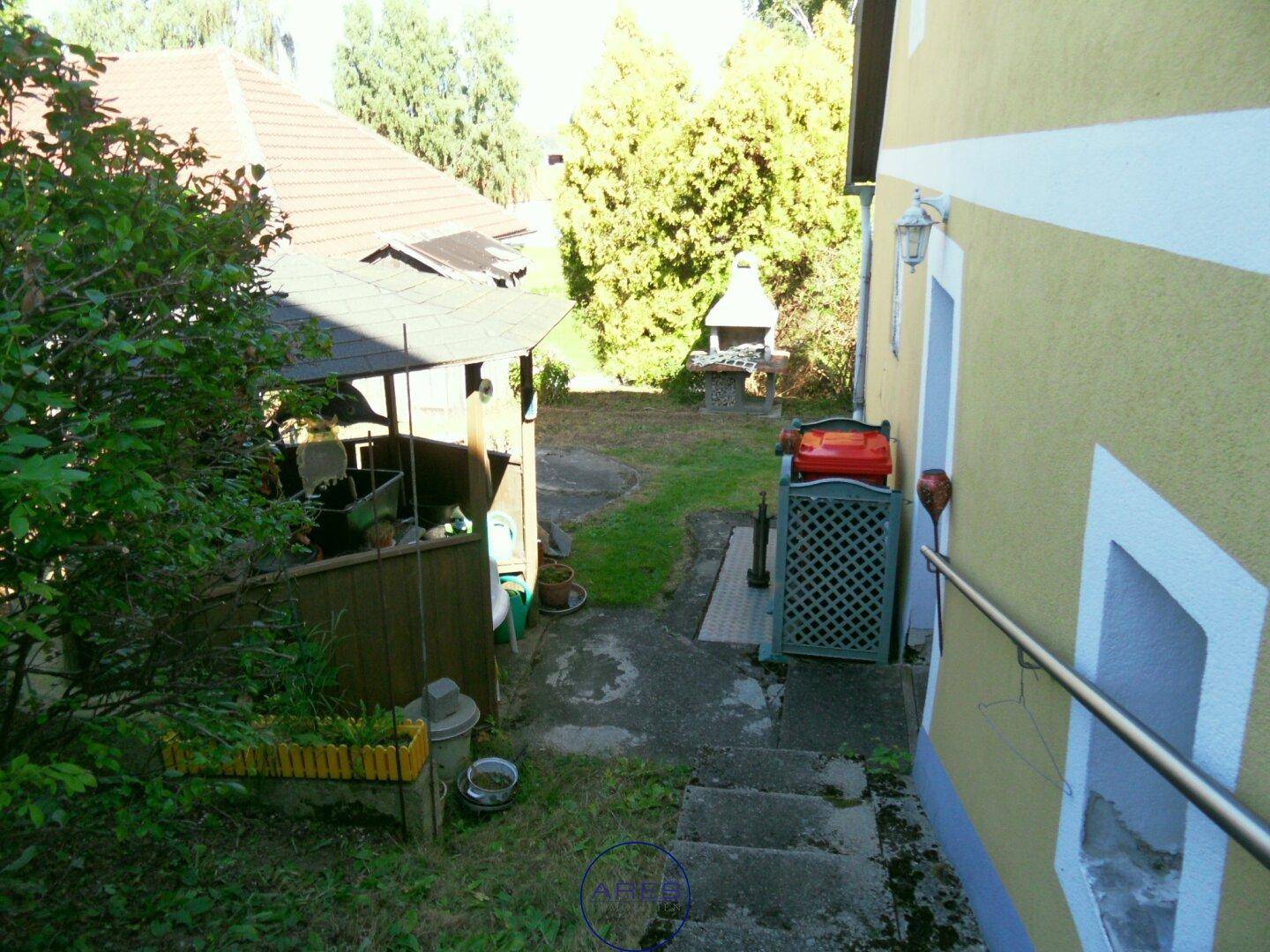 Gartenbereich mit Steintreppe, Grillplatz und überdachtem Sitzbereich neben dem Haus.