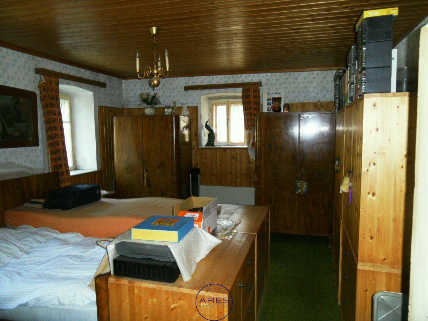 Schlafzimmer mit Holzmöbeln, gemusterter Tapete und zwei Fenstern, die Tageslicht hereinlassen.
