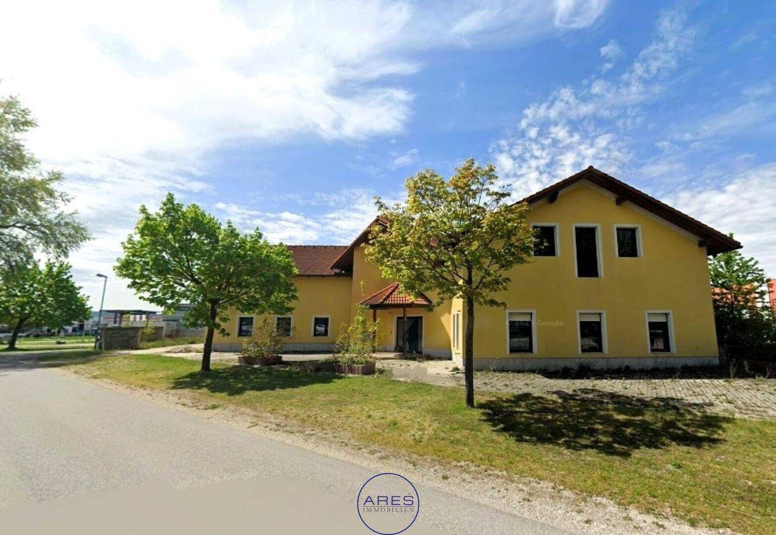 Gelbes Gebäude mit rotem Ziegeldach und grünen Bäumen an einer Straße unter blauem Himmel.