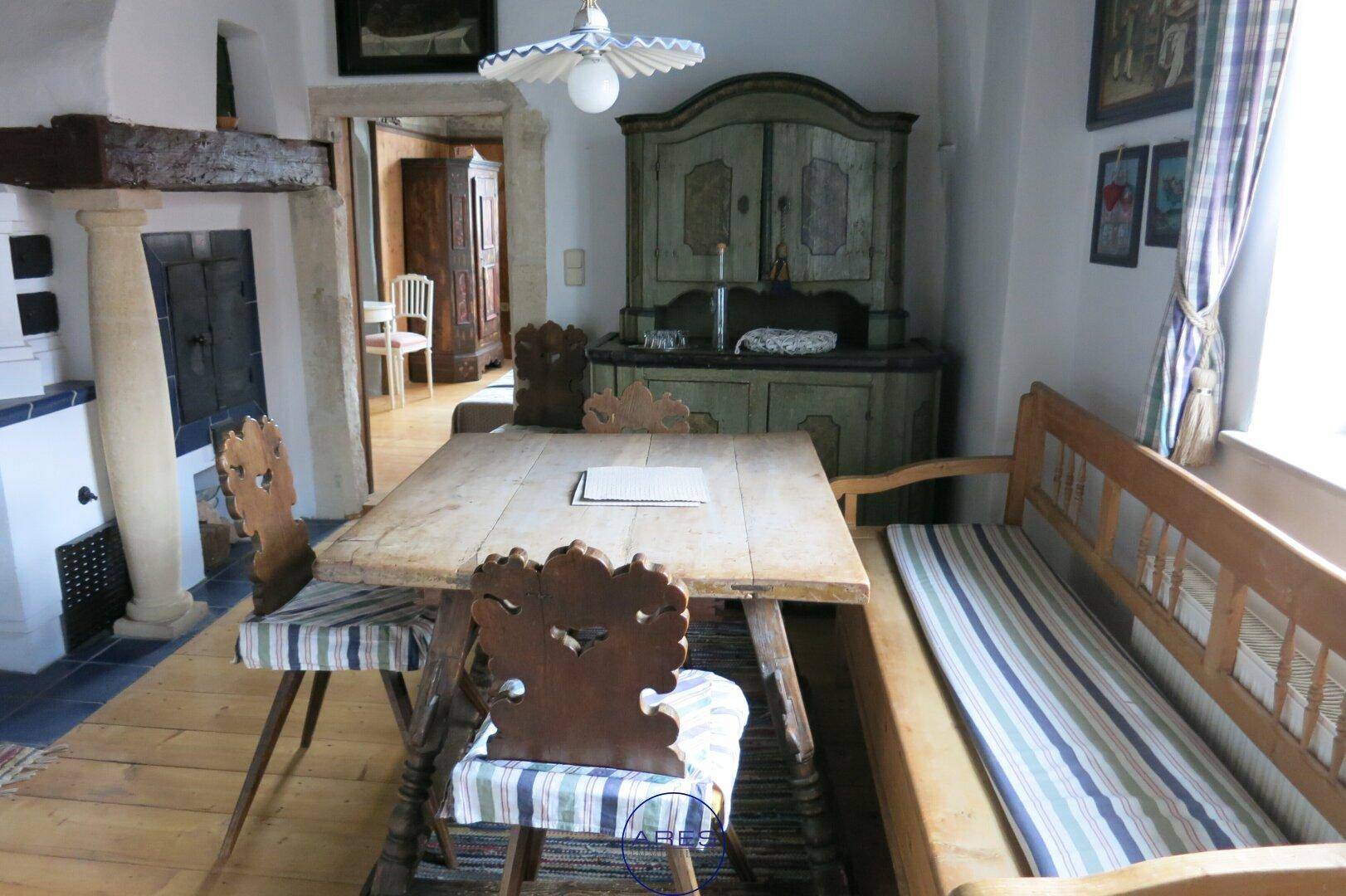 Rustikaler Essbereich mit Holztisch, Sitzbank und traditionellem Kachelofen in einem historischen Ambiente.