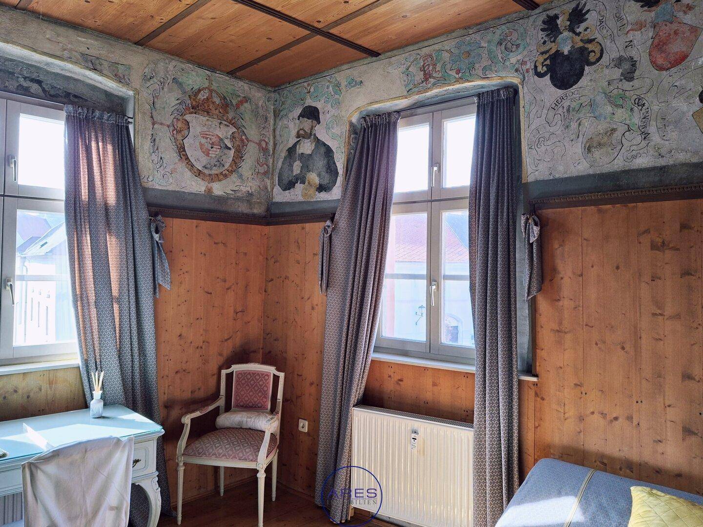 Historisches Zimmer mit bemalten Wänden, Holzvertäfelung und hohen Fenstern, die viel Licht hereinlassen.