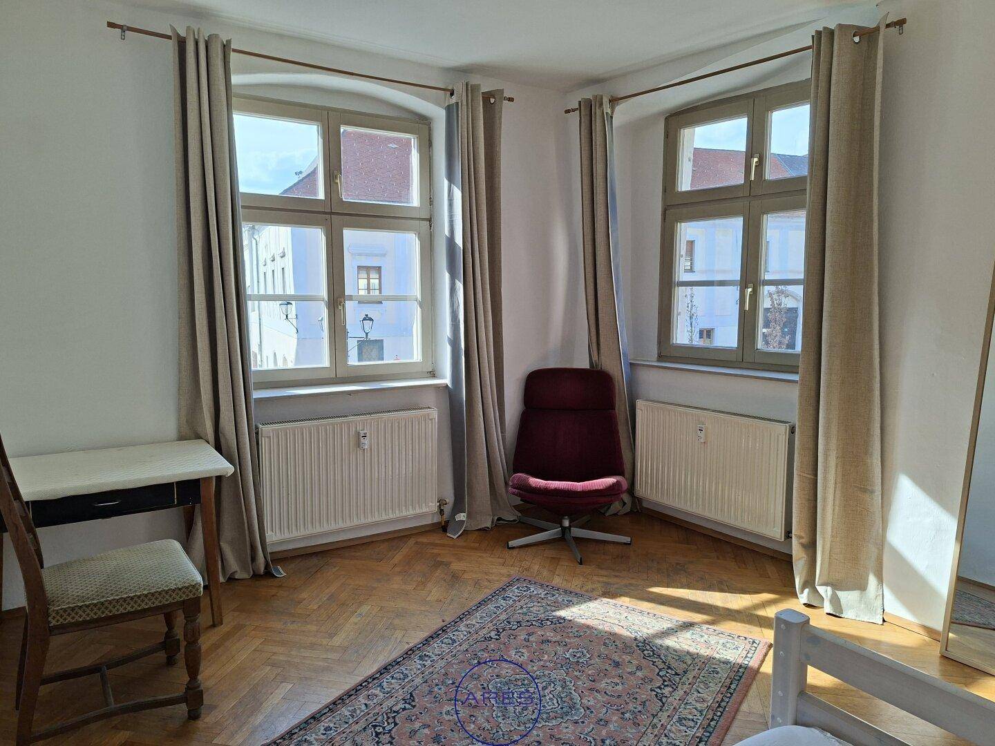Helles Zimmer mit Parkettboden, zwei großen Fenstern und Heizkörpern unter den Fenstern.
