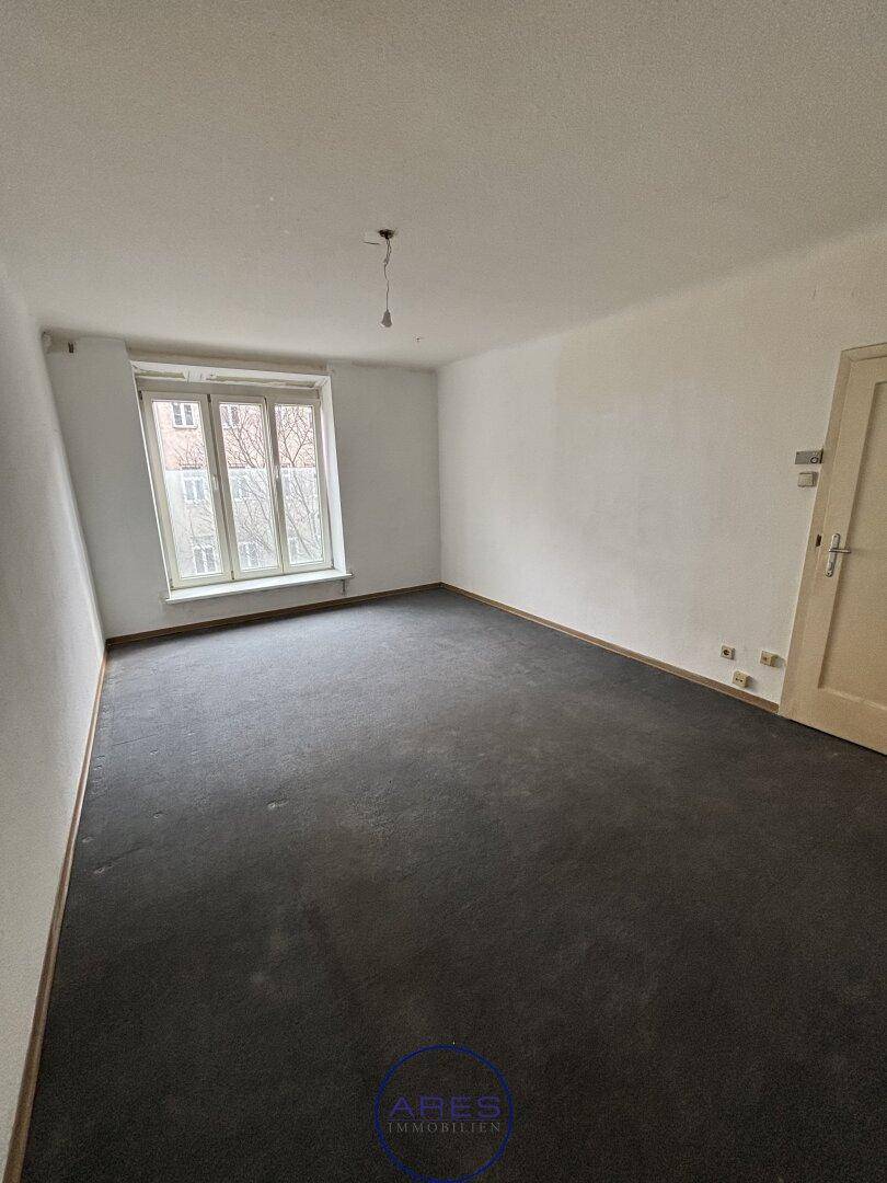 Leeres Zimmer mit dunklem Teppichboden, weißen Wänden und einem Fenster mit Blick nach draußen.