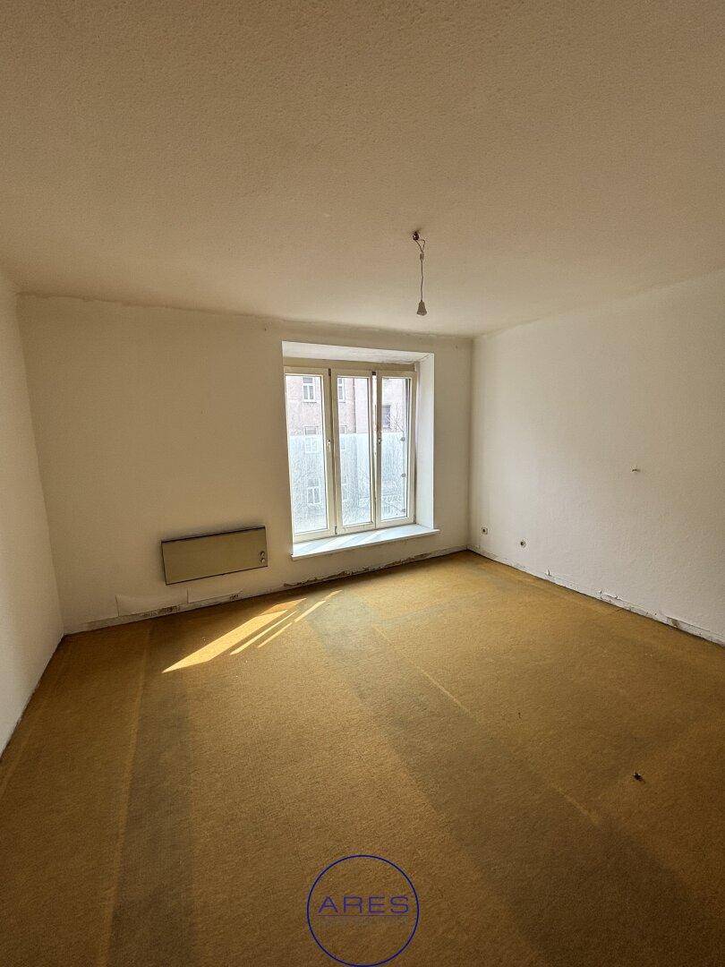Helles Zimmer mit gelbbraunem Teppichboden, weißen Wänden und einem großen Fenster.