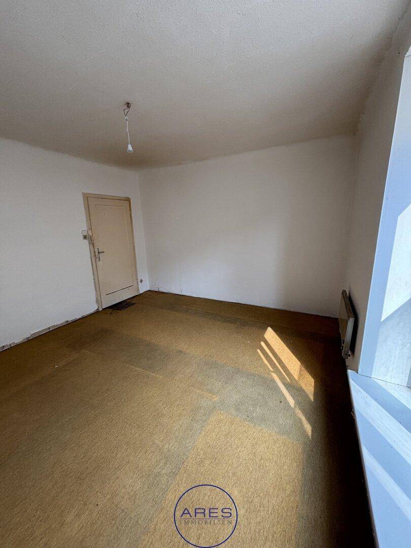 Geräumiges Zimmer mit Teppichboden, Heizkörper unter dem Fenster und einer einfachen Tür.