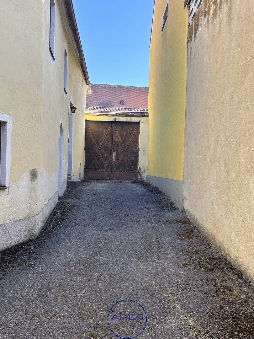 Schmaler Durchgang zwischen zwei Gebäuden, der zu einem großen Holztor führt.