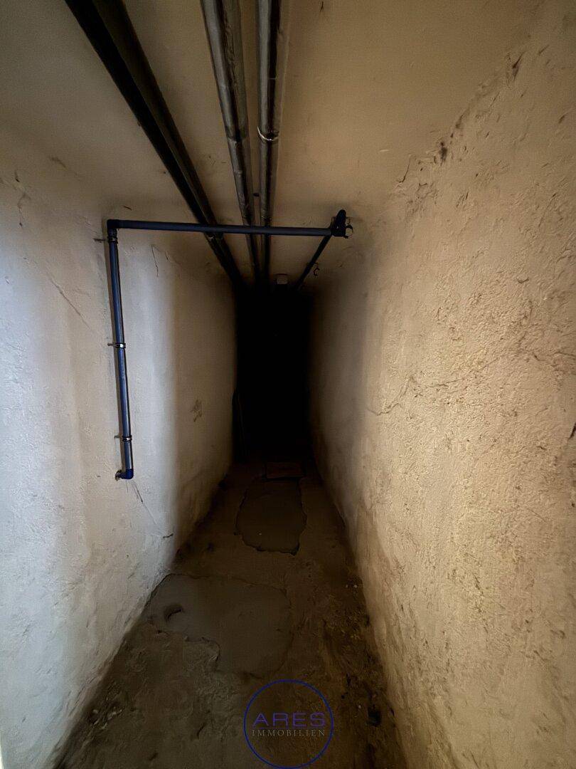 Langer, dunkler Kellergang mit Rohren an der Decke und rauen Wänden.