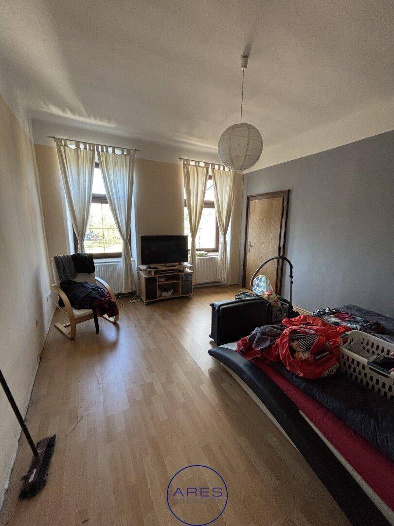 Helles Wohnzimmer mit Laminatboden, zwei Fenstern und einfacher Einrichtung.