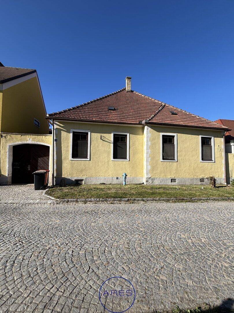 Gelbes Einfamilienhaus mit Ziegeldach und gepflastertem Vorplatz unter blauem Himmel.