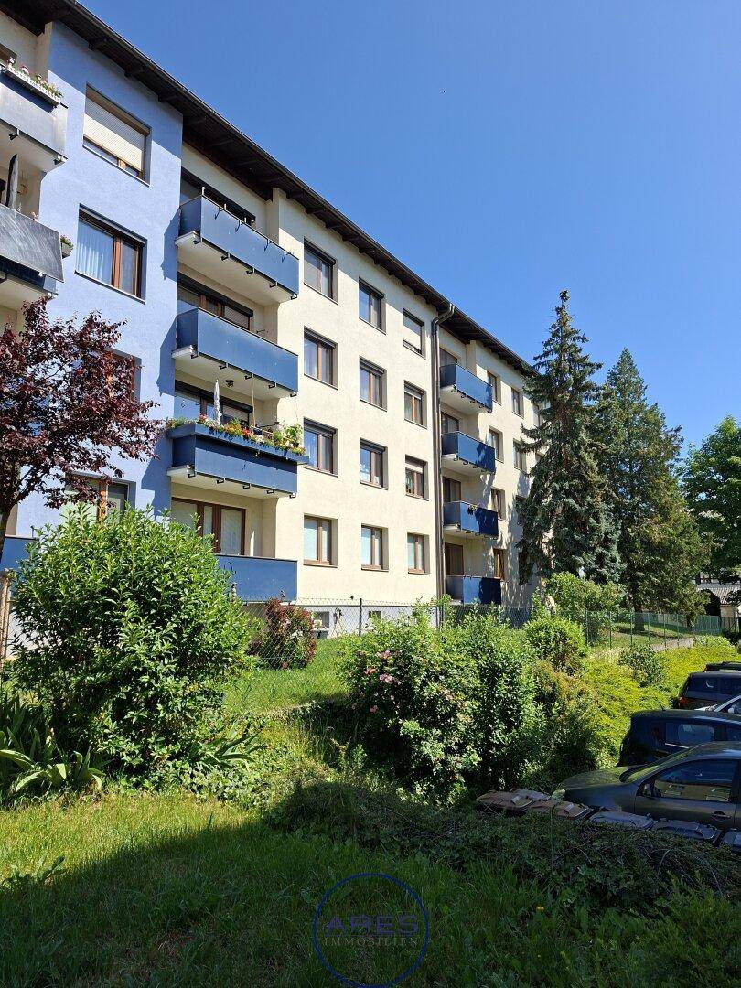 Mehrfamilienhaus mit blauen Balkonen und gepflegter Grünanlage unter blauem Himmel.