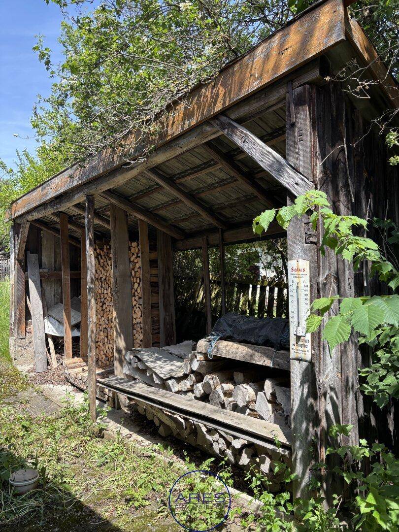 Rustikaler Holzschuppen mit gestapeltem Brennholz, umgeben von Bäumen und Sträuchern.