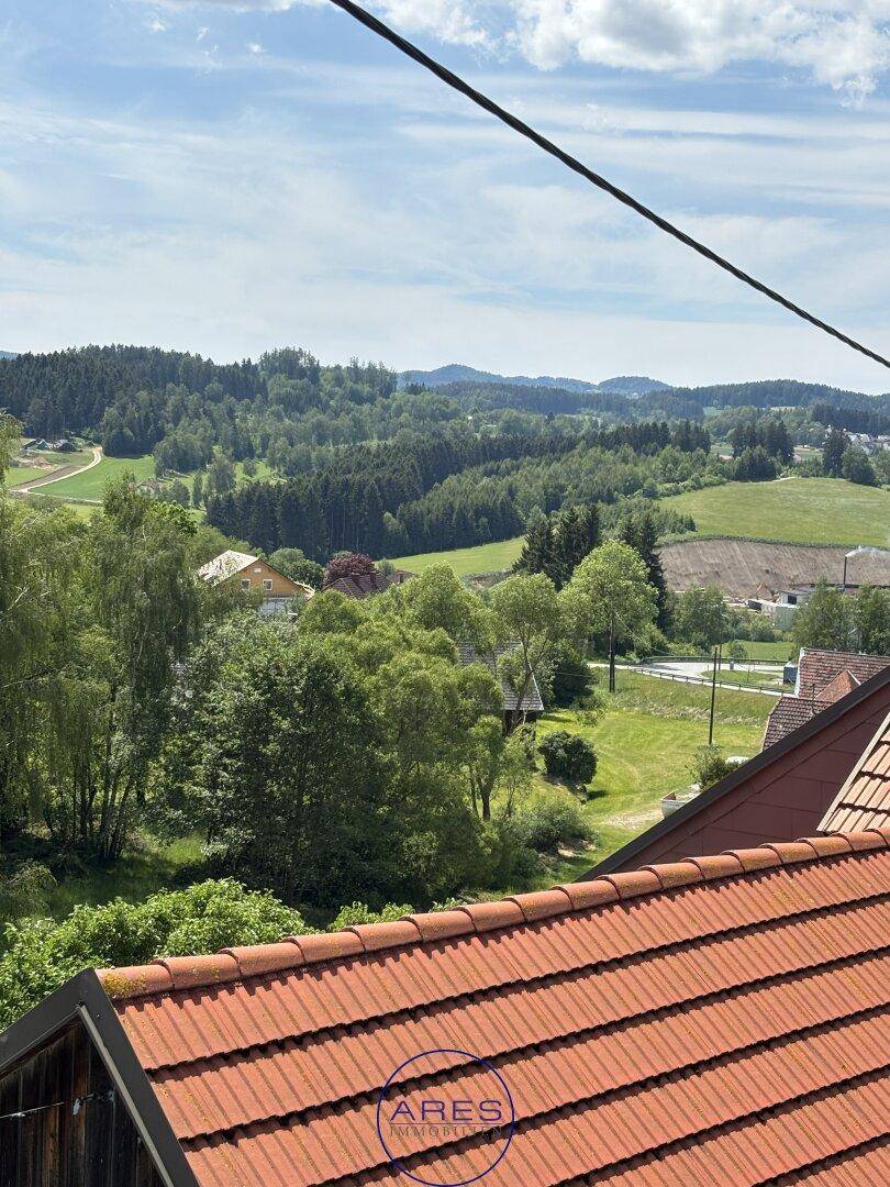 Malerischer Blick über Ziegeldächer auf eine weite, grüne Hügellandschaft unter blauem Himmel.