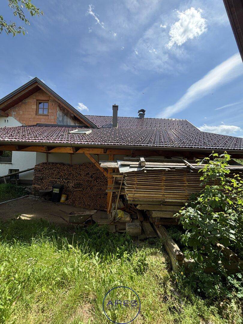 Seitenansicht des Hauses mit Holzstapeln und gelagertem Bauholz unter einem schützenden Unterstand.