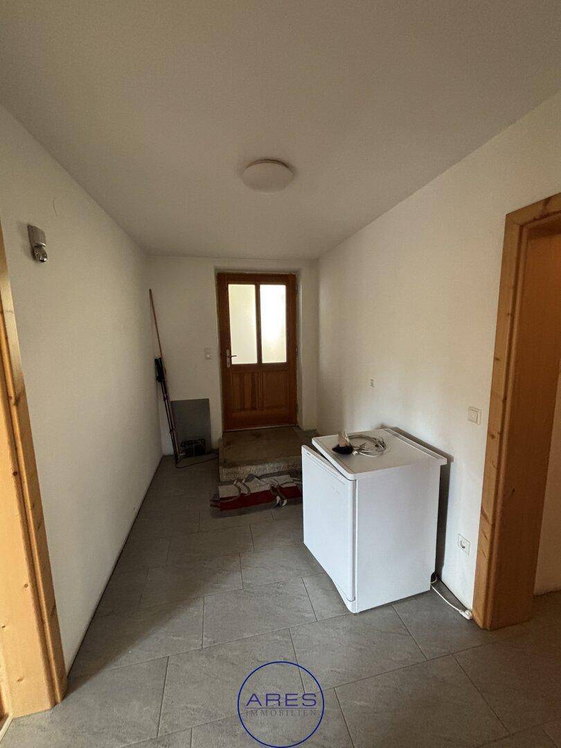 Heller Flur mit Fliesenboden und einer Holztür am Ende, ein kleiner Kühlschrank steht bereit.