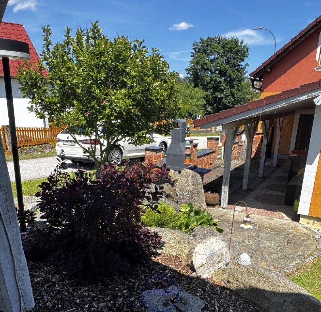 Sonniger Gartenbereich mit gepflastertem Weg, Steinen, Pflanzen und überdachtem Grillplatz.