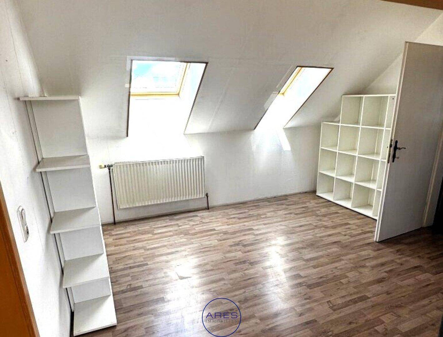 Helles Zimmer im Dachgeschoss mit Laminatboden, Regalen und zwei Dachfenstern.