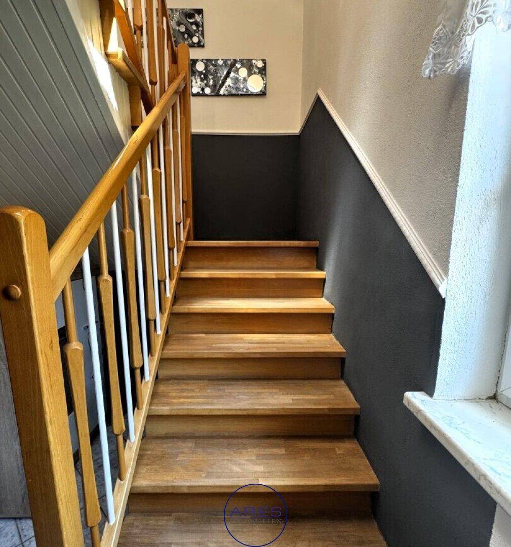 Holztreppe mit Geländer und zweifarbiger Wandgestaltung, die in das Obergeschoss führt.