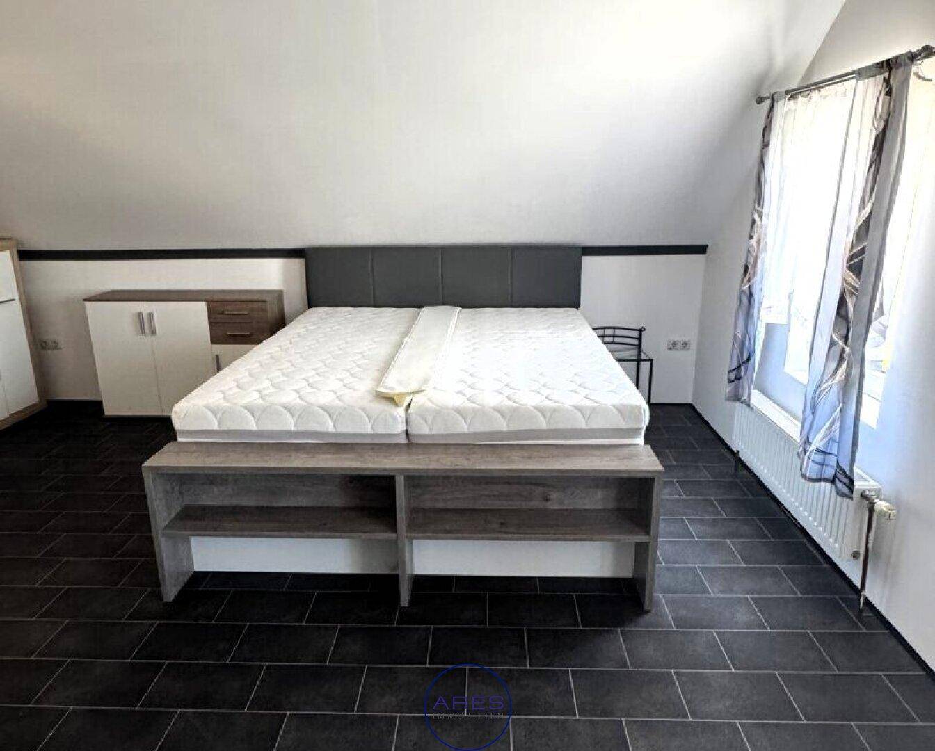 Helles Schlafzimmer im Dachgeschoss mit Doppelbett, dunklen Fliesen und Dachschrägen.
