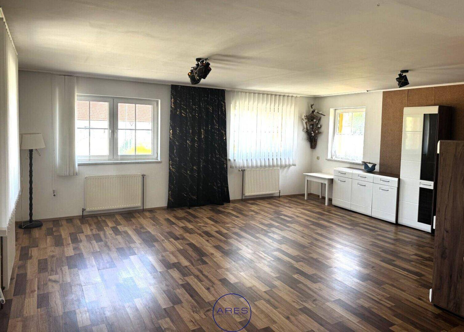 Geräumiges Wohnzimmer mit Laminatboden, großen Fenstern und moderner Beleuchtung.