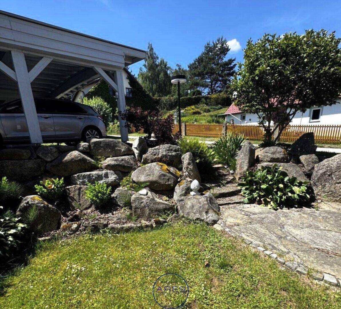 Weitläufiger Gartenbereich mit Steinen, Rasen und einem überdachten Stellplatz für Fahrzeuge.