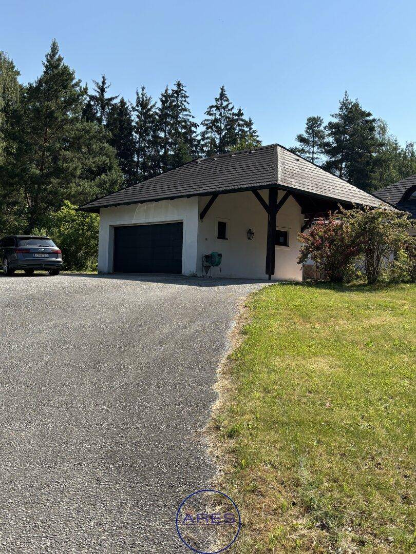 Separate Doppelgarage mit Carport und Zufahrt, umgeben von grüner Natur unter blauem Himmel.