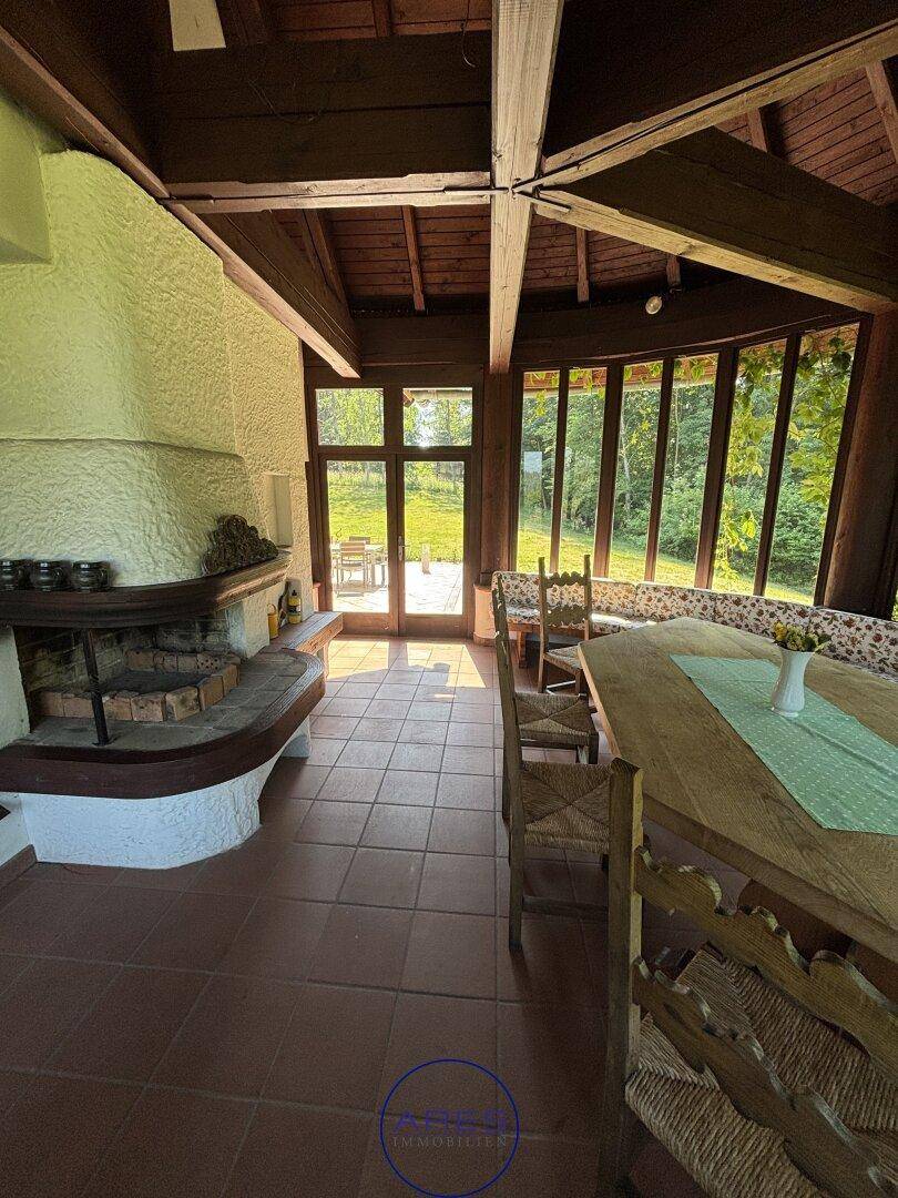 Gemütlicher Wintergarten mit Kamin, Holzbalken und großen Fenstern mit Blick in den Garten.