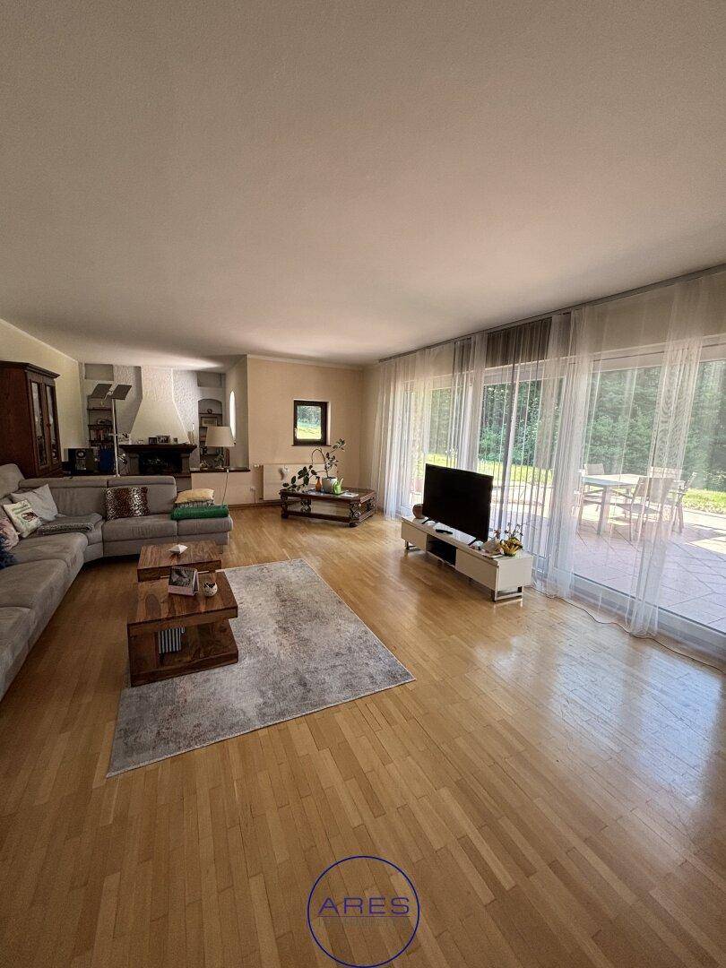 Lichtdurchflutetes Wohnzimmer mit Holzboden, großem Sofa und bodentiefen Fenstern mit Blick ins Grüne.