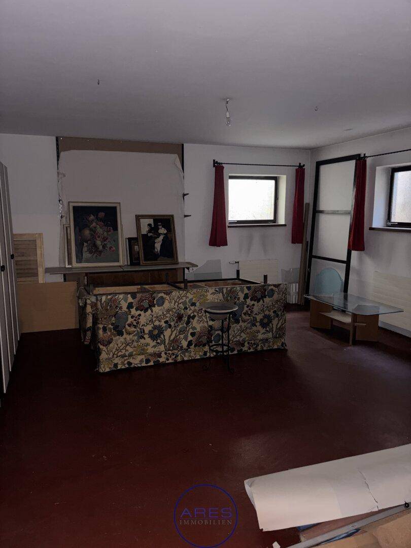 Geräumiger Wohnraum mit dunklem Boden, weißen Wänden und zwei Fenstern mit roten Vorhängen.