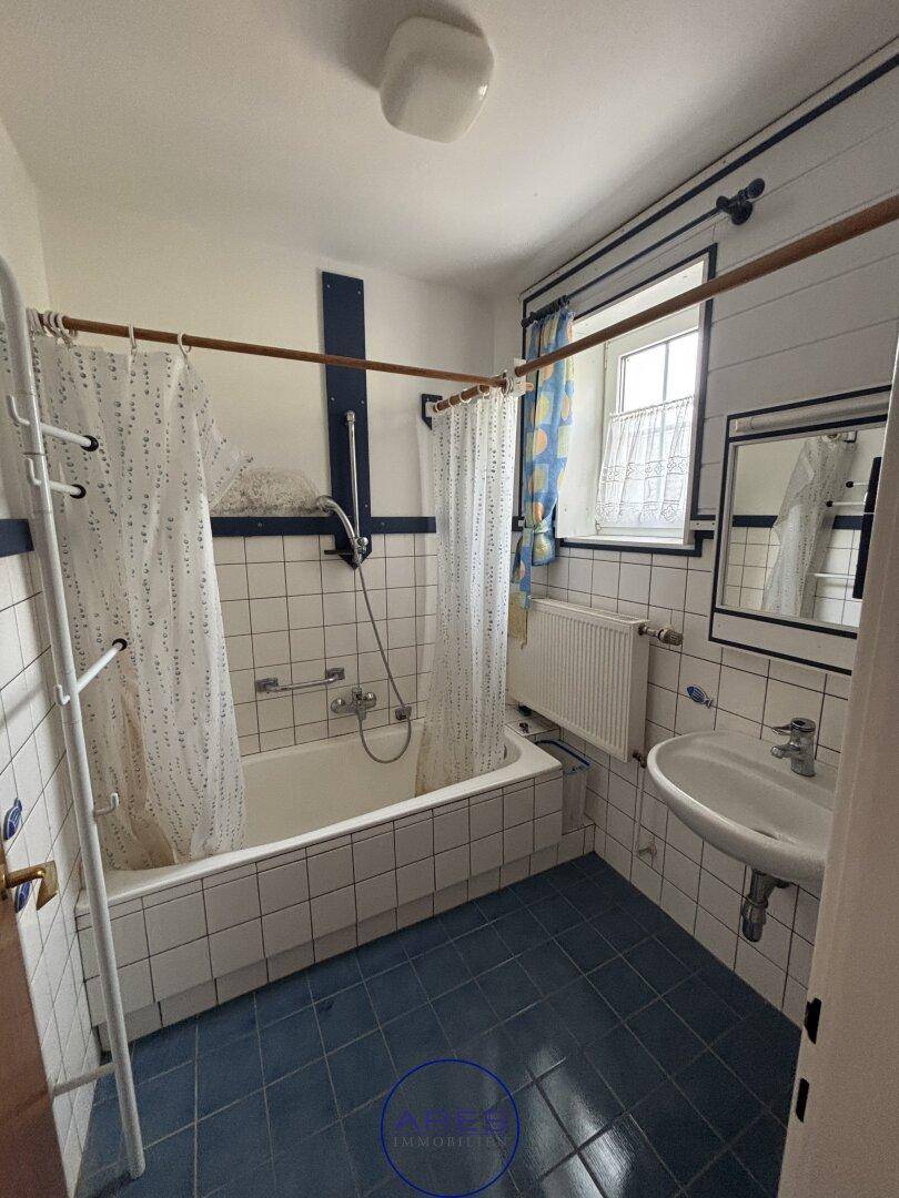 Badezimmer mit Badewanne, blauen Bodenfliesen und weißen Wandfliesen im 80er-Jahre-Stil.
