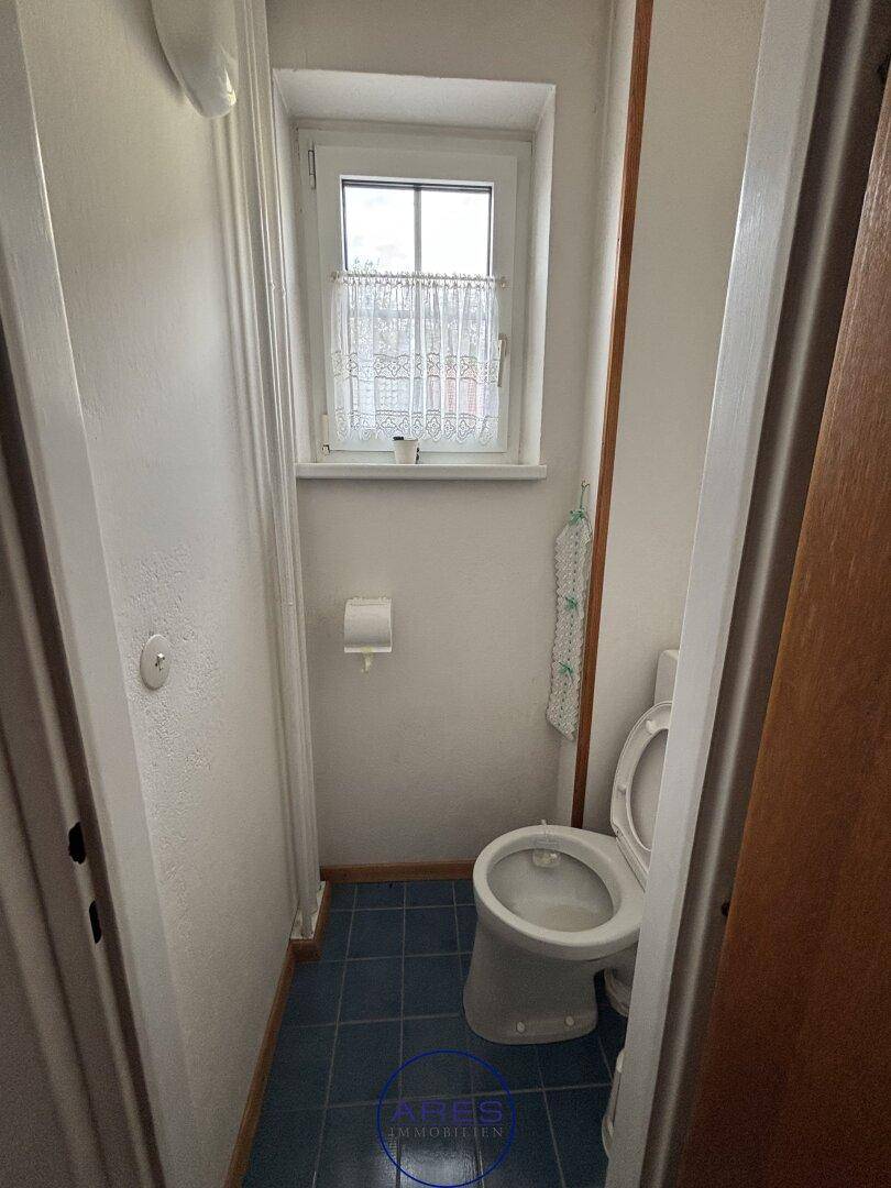 Separate Toilette mit Fenster, weißen Wänden und blauen Bodenfliesen.