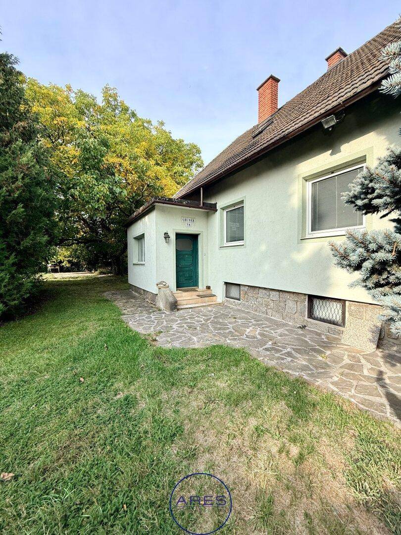 Seitenansicht des Hauses mit grünem Eingangsbereich und kleiner Erweiterung, umgeben von Rasen und Bäumen.